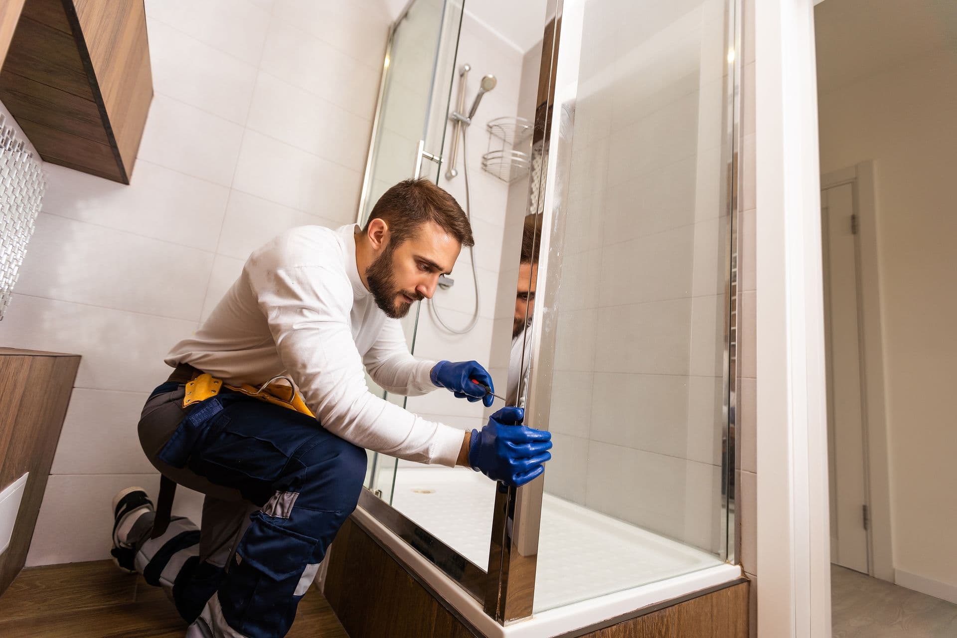 A man in work attire and blue gloves kneels to install a glass shower door in a modern bathroom, focusing intently on aligning it accurately.